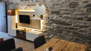 A rustic interior with a stone wall, a wooden ceiling, a flat-screen TV, a dining table with a plate of cookies, and a hanging light fixture. - Castas D'ouro Guest House II (Casal de Loivos)