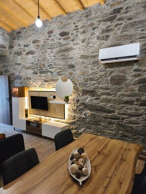 A rustic interior with a stone wall, a wooden ceiling, a flat-screen TV, a dining table with a plate of cookies, and a hanging light fixture. - Castas D'ouro Guest House II (Casal de Loivos)