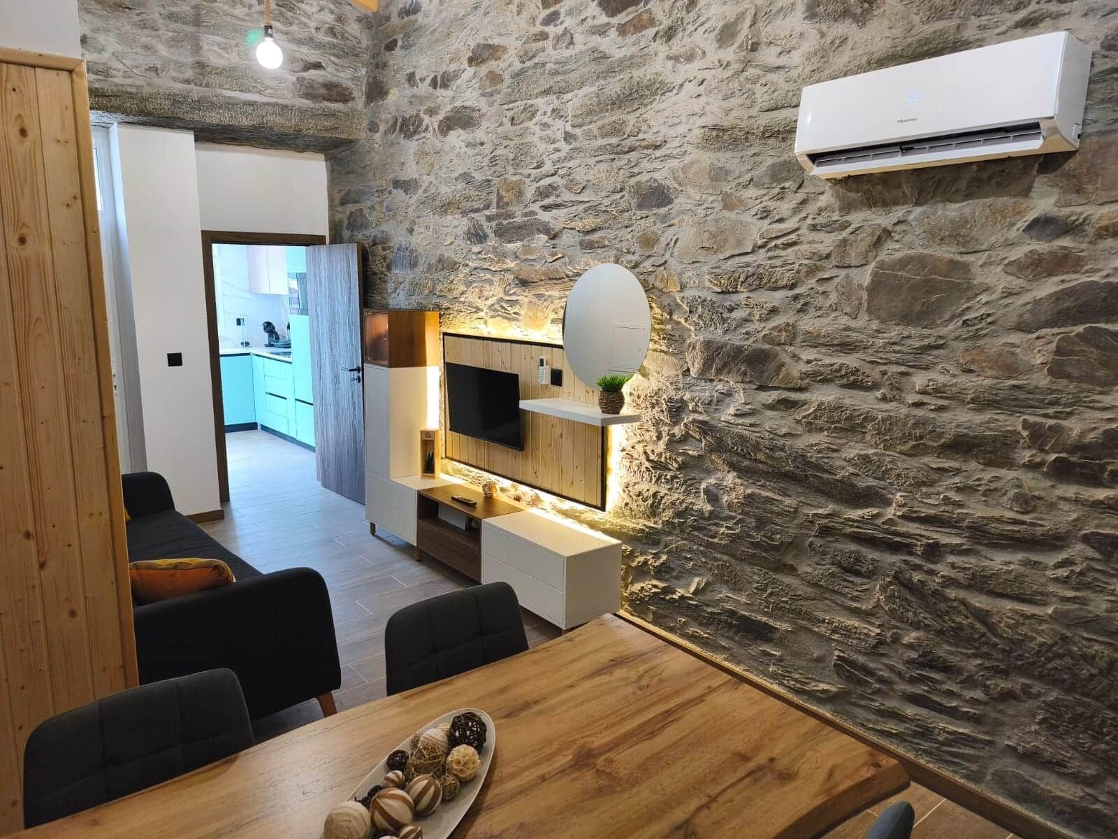 A modern living space with a stone wall, a flat-screen TV, and a dining area with a wooden table and chairs.