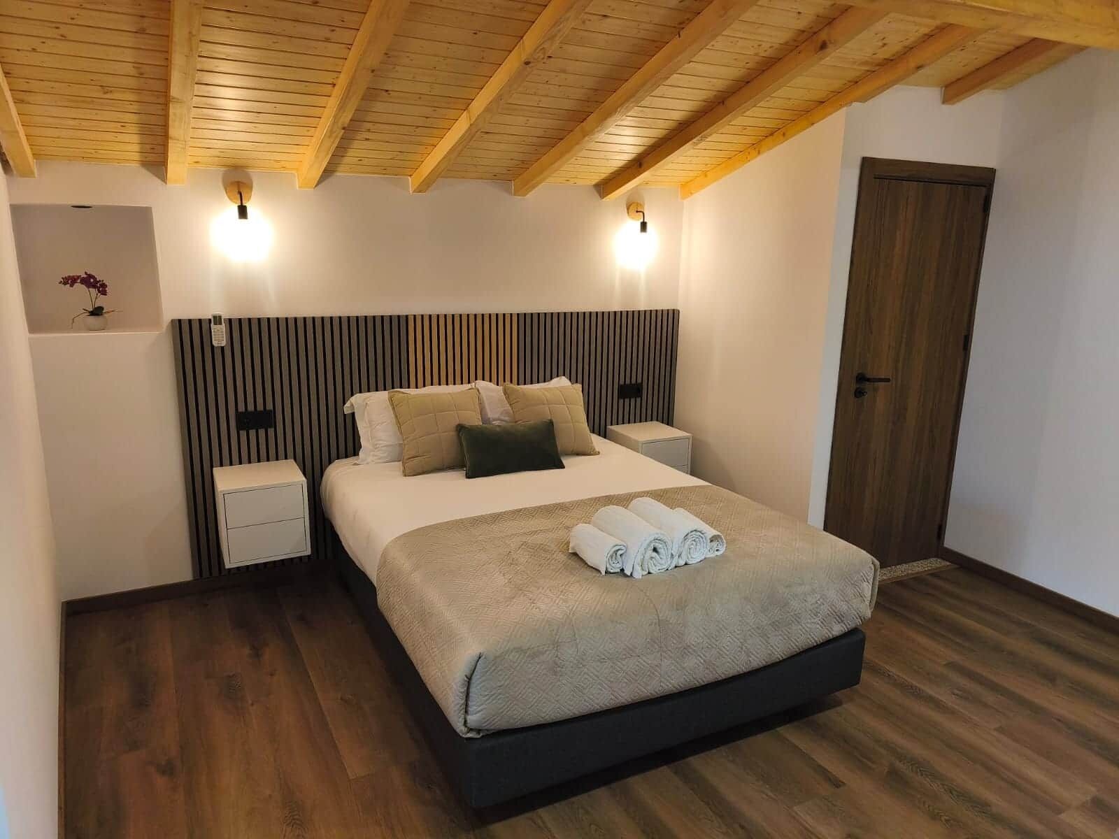 A bedroom with a wooden ceiling, a bed with a headboard, bedside tables, and a door.