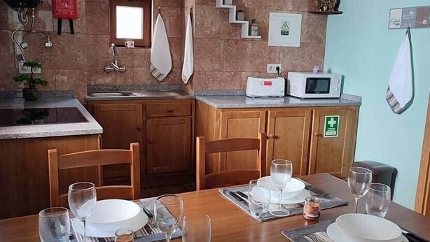 Ein Essbereich mit einem Holztisch, gedeckt für zwei Personen, eine Küche mit Mikrowelle und Spüle sowie eine Uhr an der Wand.