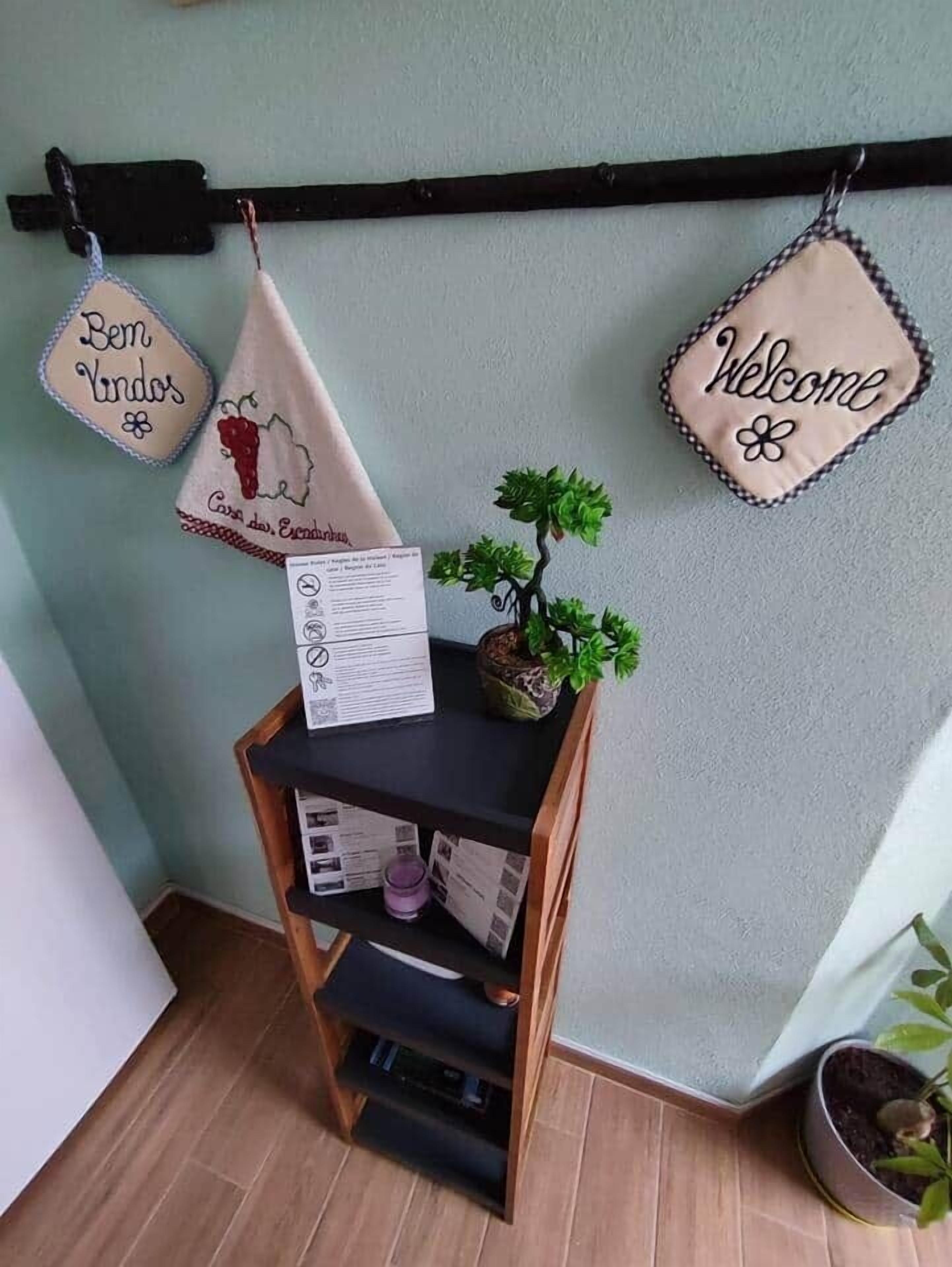 Un rincón con un estante de madera que sostiene una planta pequeña en maceta y un menú, flanqueado por dos letreros en la pared que dicen 'Bem Vindos' y 'Welcome'.