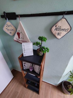 Un rincón con un estante de madera que sostiene una planta pequeña en maceta y un menú, flanqueado por dos letreros en la pared que dicen 'Bem Vindos' y 'Welcome'.