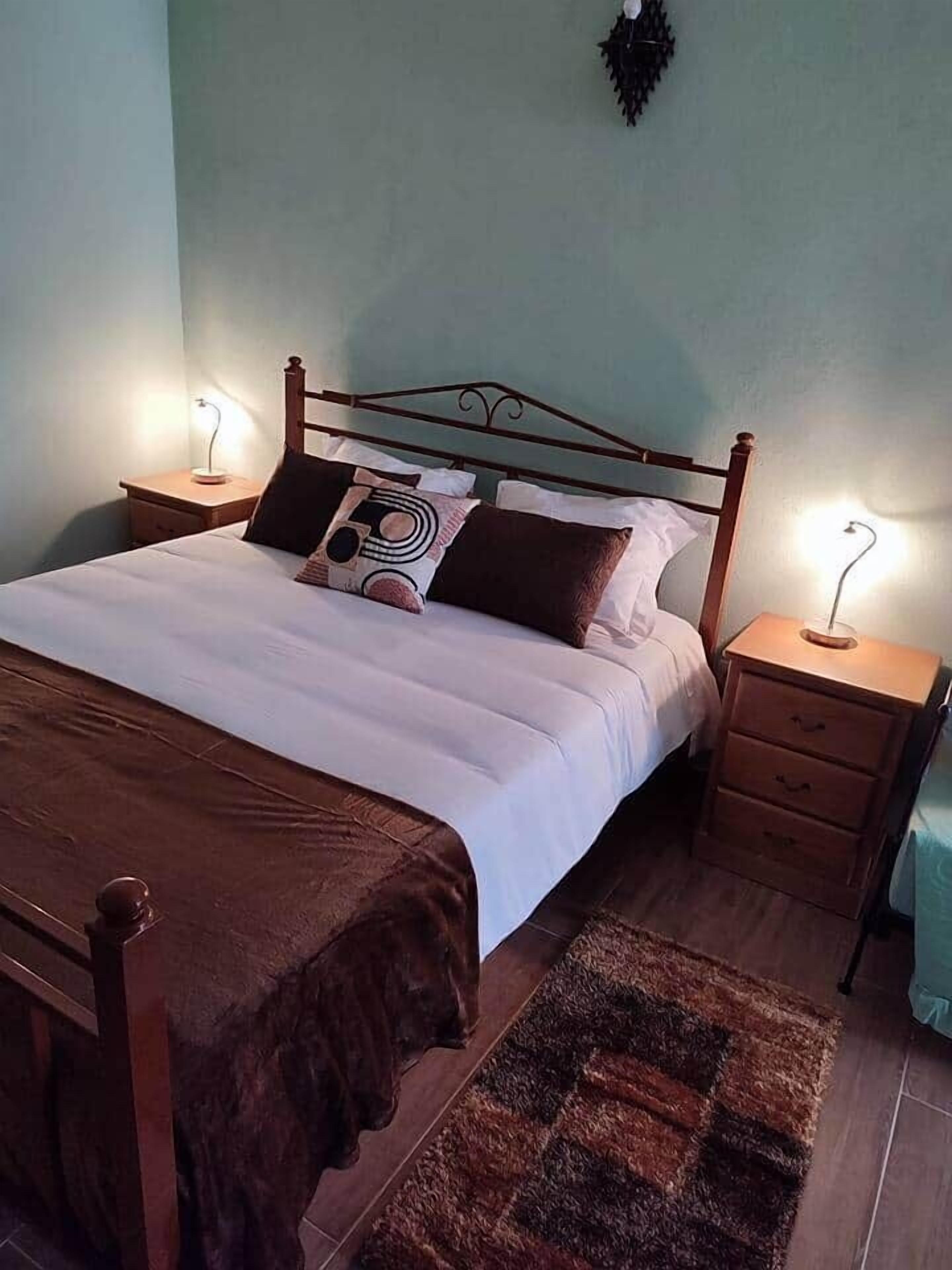 A single bed with a wooden frame, two bedside tables with lamps, a patterned rug, and a wall-mounted light fixture.