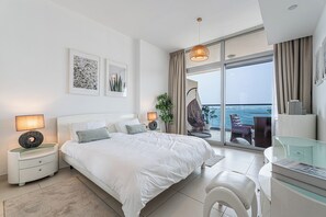 Een moderne slaapkamer met een groot bed, nachtkastjes, een bureau en uitzicht op zee door een schuifdeur.