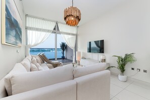 A modern living room with a large sofa, a flat-screen TV, and a view of the sea through large windows. - Luxury 1-bedroom apartment with direct beach access (DUBAI)