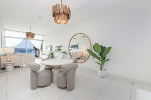 A modern living room with a round dining table, grey upholstered chairs, a large mirror, and a view of the sea through large windows. - Luxury 1-bedroom apartment with direct beach access (DUBAI)