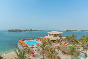 A pool area with a clear view of the sea, surrounded by palm trees and orange umbrellas. - Luxury 1-bedroom apartment with direct beach access (DUBAI)