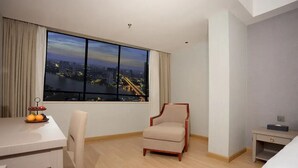 Ein Hotelzimmer mit großem Fenster und Blick auf die Stadt, einem Esstisch mit Früchten, einem Stuhl und einem Nachttisch.
