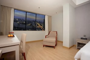 Habitación de hotel con un ventanal grande que ofrece vistas a la ciudad, una mesa de comedor con fruta, una silla y una mesita de noche.