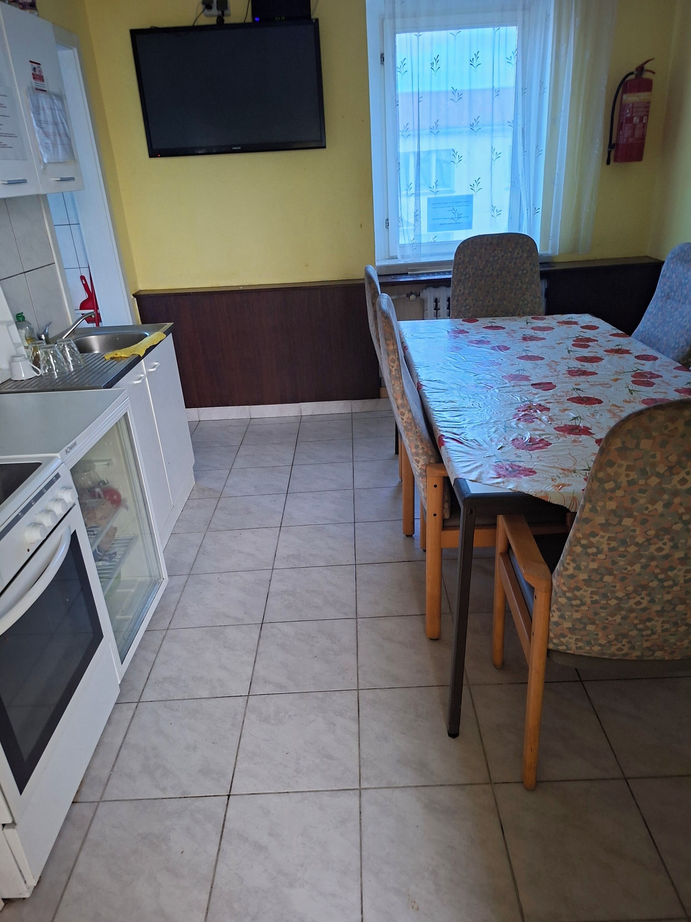 Eine kompakte Küche mit Einbauofen und kleinem Kühlschrank, ein Esstisch mit Blumenmuster bedecktem Tischläufer und ein Fenster mit Vorhängen.