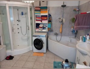 A bathroom with a glass-enclosed shower, a washing machine, a bathtub, and a shelf with folded towels. - Vrbo Property (Wanzleben-Börde)