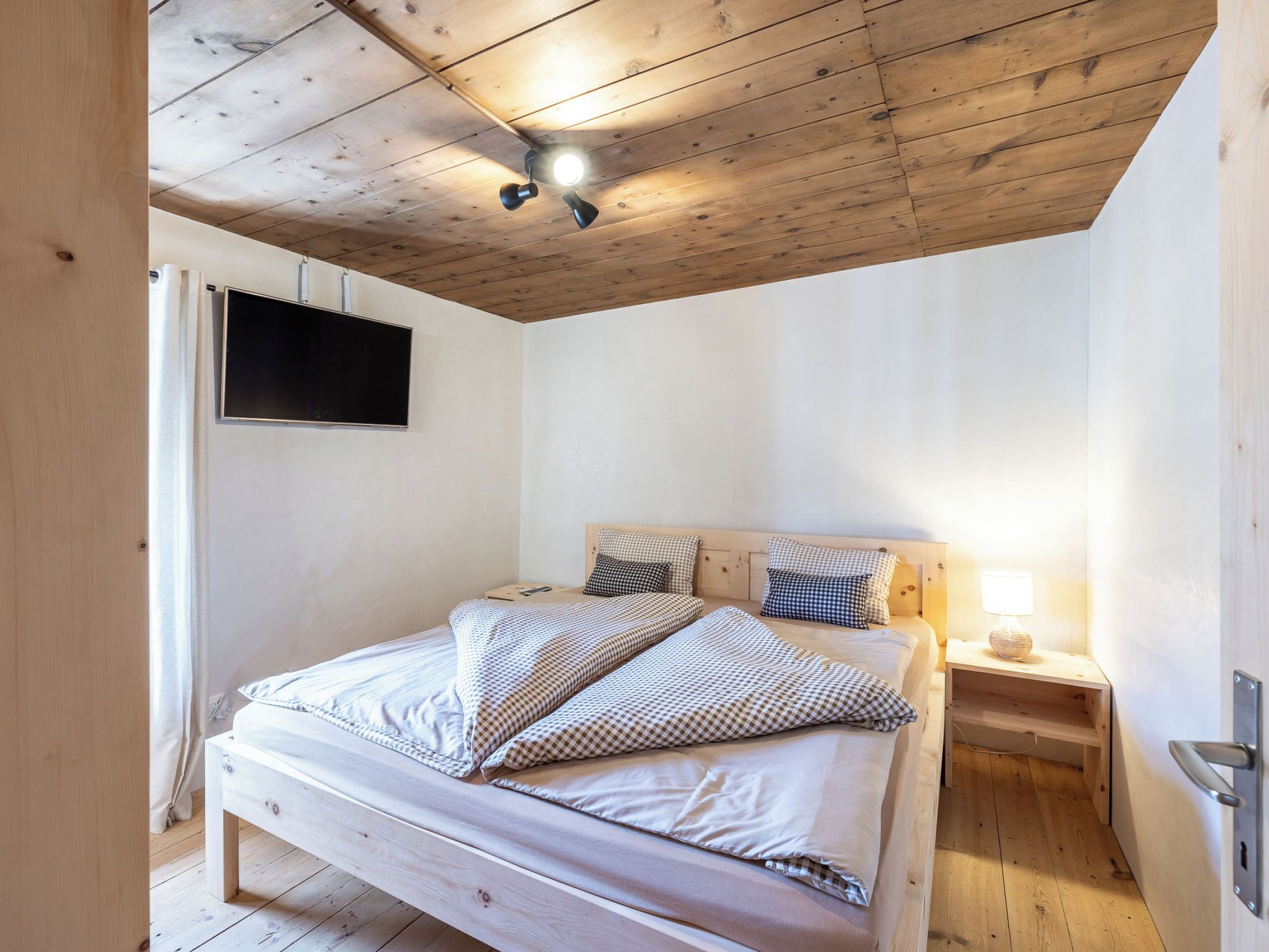 Un dormitorio con techo de madera, una cama con ropa de cama a cuadros, un televisor de pantalla plana y una lámpara de noche.