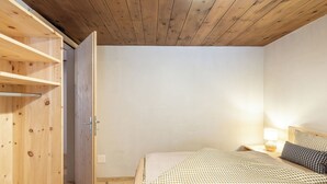 A bedroom with a wooden wardrobe, a bed with checkered bedding, a bedside lamp, and a wooden ceiling.