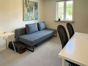 A room with a grey sofa, a small table, a chair, and a window with a plant on the sill.