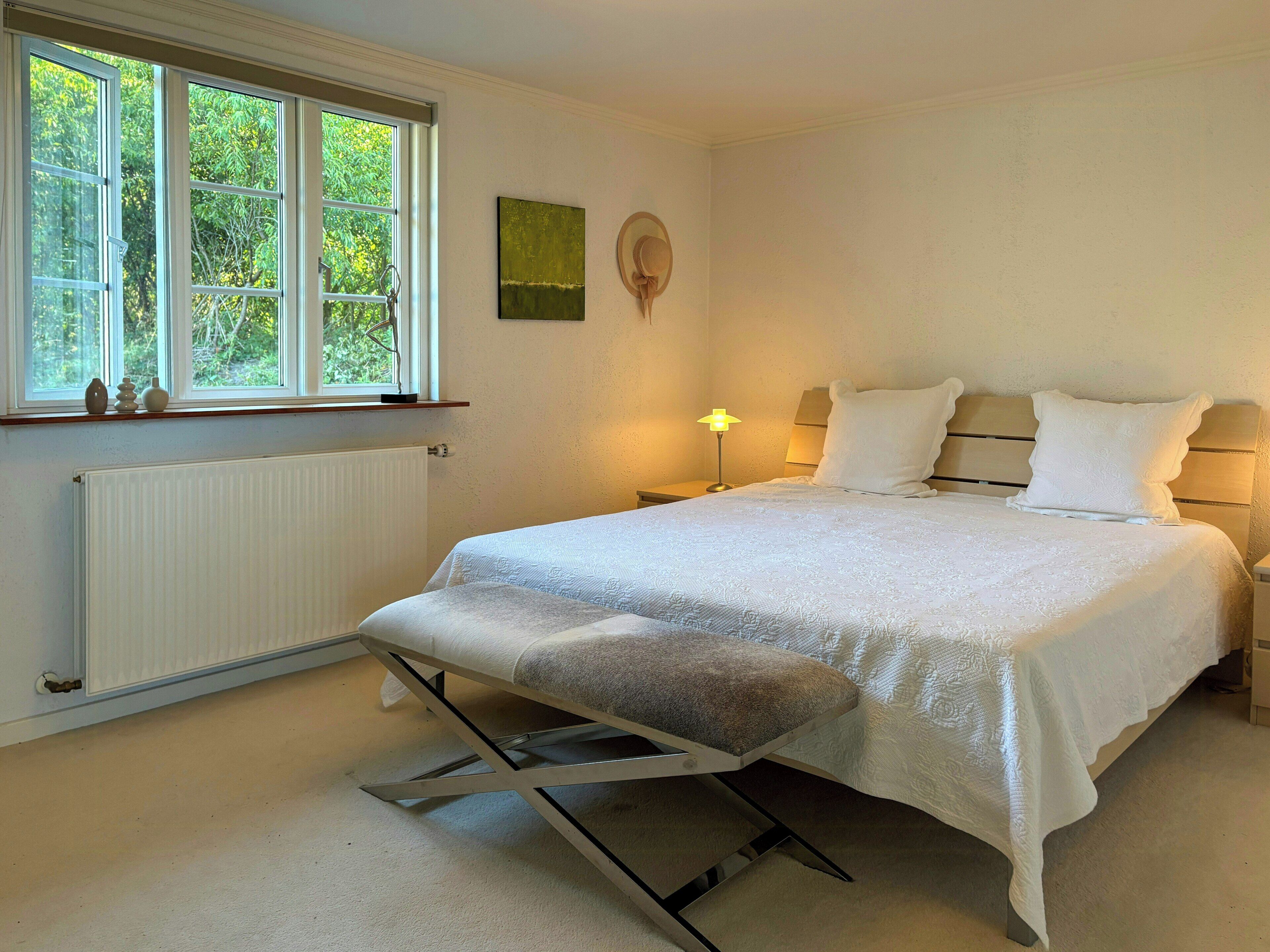 A bedroom with a bed, bedside lamps, a window with a view of greenery, a radiator, and a small table with decorative items.