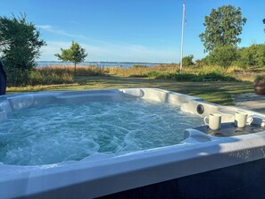 Un jacuzzi con dos tazas a un lado, rodeado de césped y árboles, con un asta de bandera y una masa de agua al fondo.