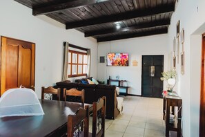 Un saló amb sostre de fusta, un televisor fixat a la paret, un sofà, una taula de dinar amb cadires i una porta.
