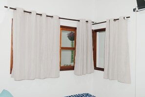 Ein Zimmer mit Fenster, weißen Vorhängen, einem Holzrahmen und einer zudeckenden, blau gemusterten Bettdecke.