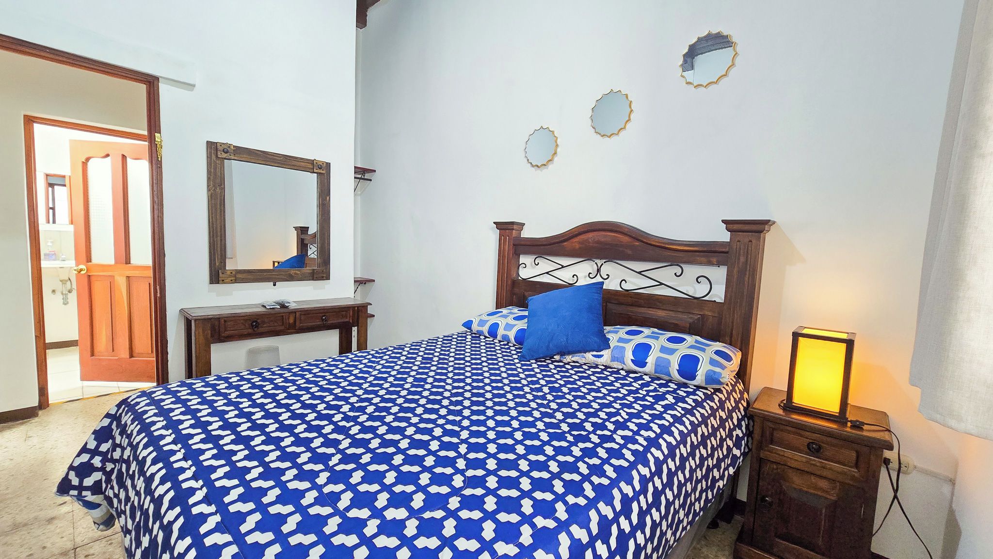 Quarto com cama, mesa de cabeceira, abajur, espelho e porta.