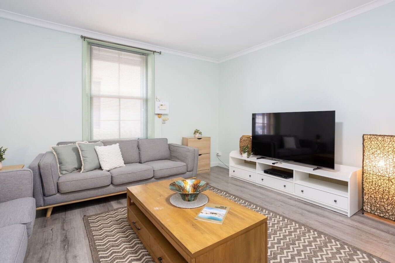 A modern living room with a grey sofa, a wooden coffee table, a flat-screen TV on a white stand, and a patterned rug.