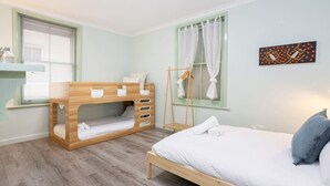 A bedroom with a bunk bed, a single bed, a window with curtains, a towel rack, and a wall art piece. - Heart of Freo - Charming Heritage Stay (Fremantle)