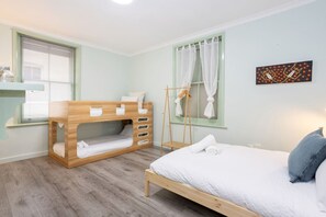 A bedroom with a bunk bed, a single bed, a window with curtains, a towel rack, and a wall art piece. - Heart of Freo - Charming Heritage Stay (Fremantle)