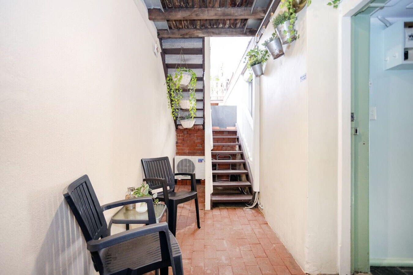 A narrow outdoor corridor with a brick floor, a wooden staircase, and a small table with chairs.