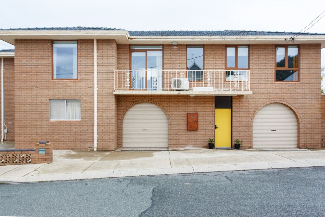 A two-story brick building with a yellow door, two garage doors, and a small balcony with a white railing.