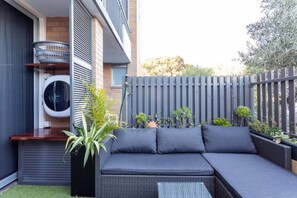 Zona d'estar exterior moderna amb un sofà integrat i una taula petita, envoltada de plantes en testos i una tanca gris.
