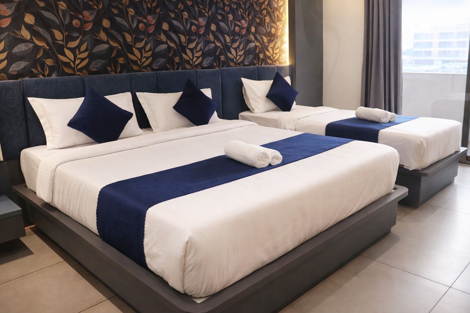 Una habitación de hotel con dos camas, cada una con ropa de cama blanca y detalles azules, un cabecero oscuro y una pared decorativa con un diseño de hojas.