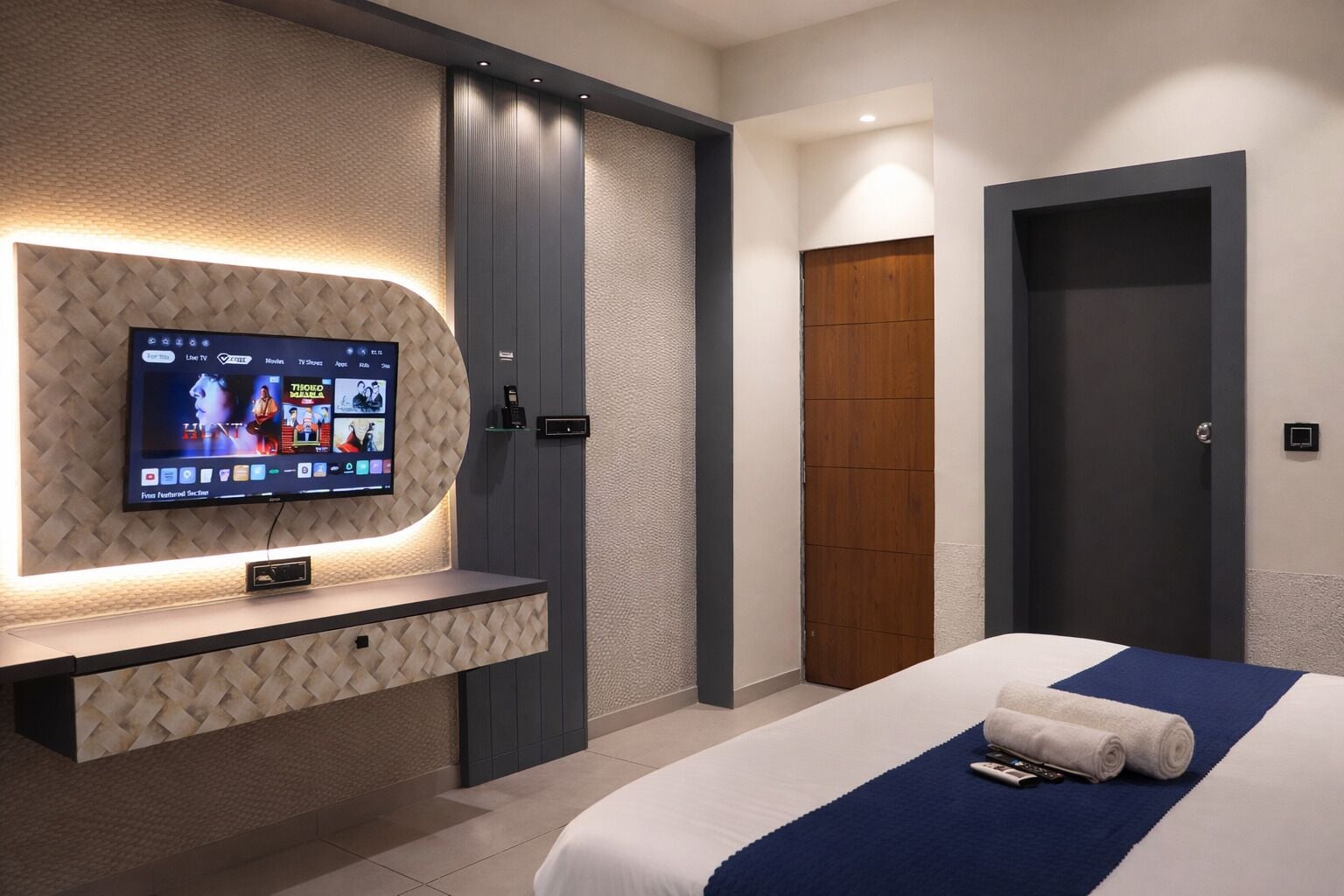 Habitación de hotel con televisor montado en una pared texturizada, cama con detalles en azul y puerta de madera.