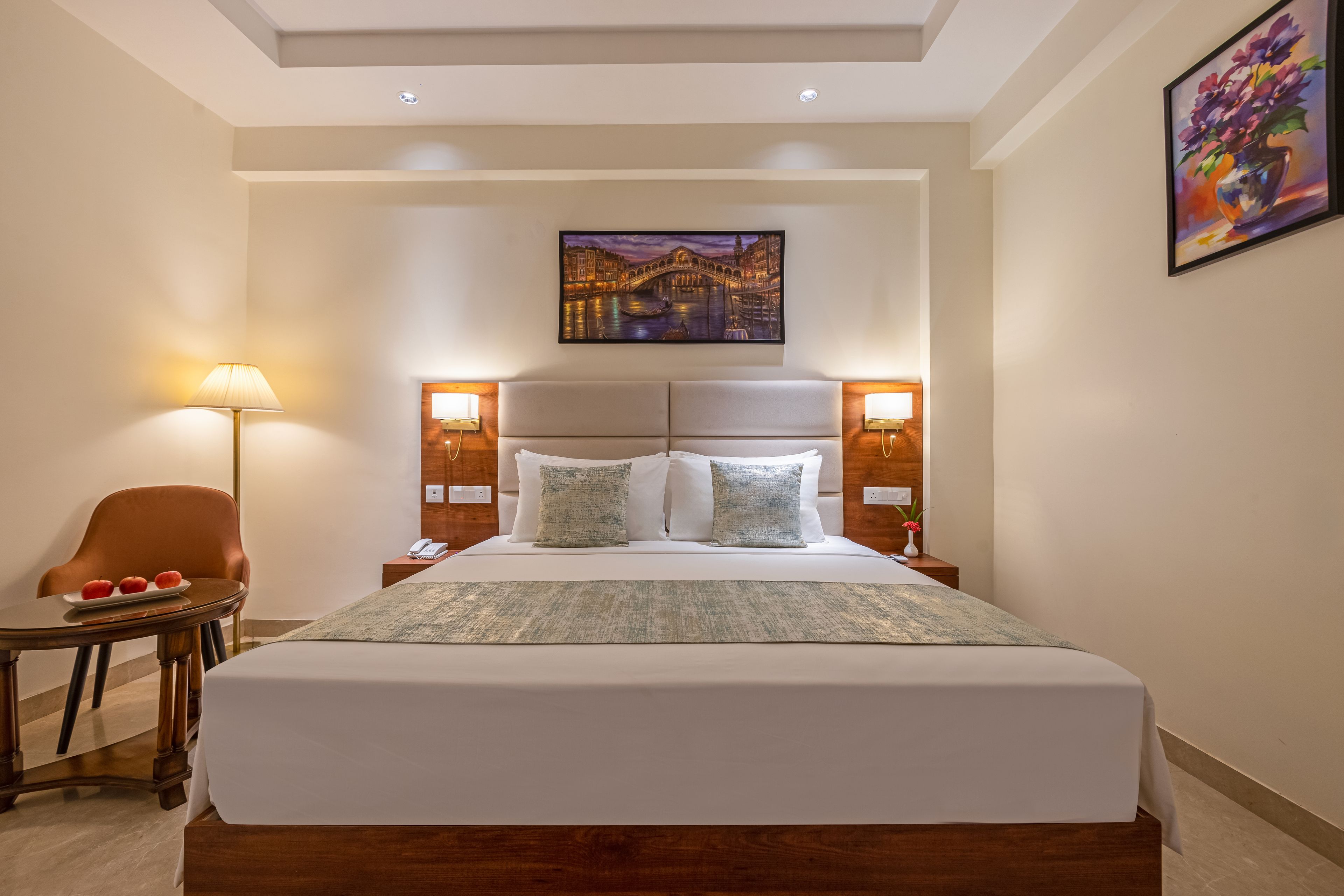 Una camera d'albergo con un letto grande, abat-jour, una sedia, un tavolino con delle mele e un quadro appeso al muro.
