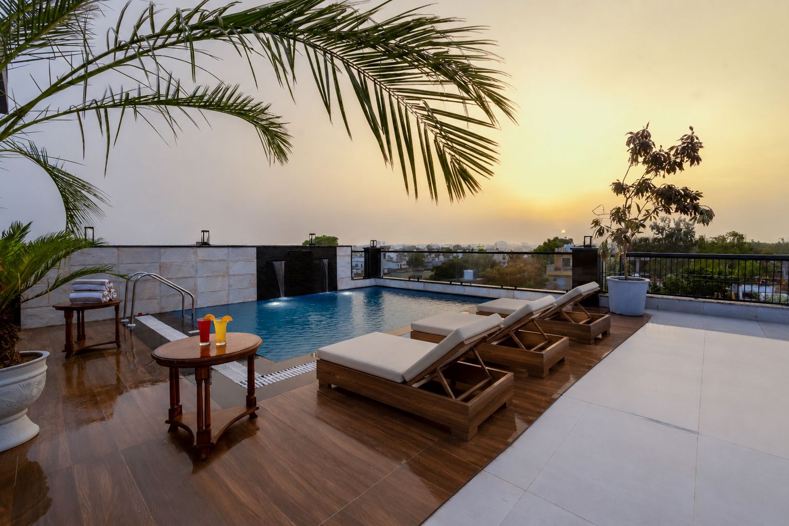 A rooftop pool area with lounge chairs, a small table with drinks, and a waterfall feature.