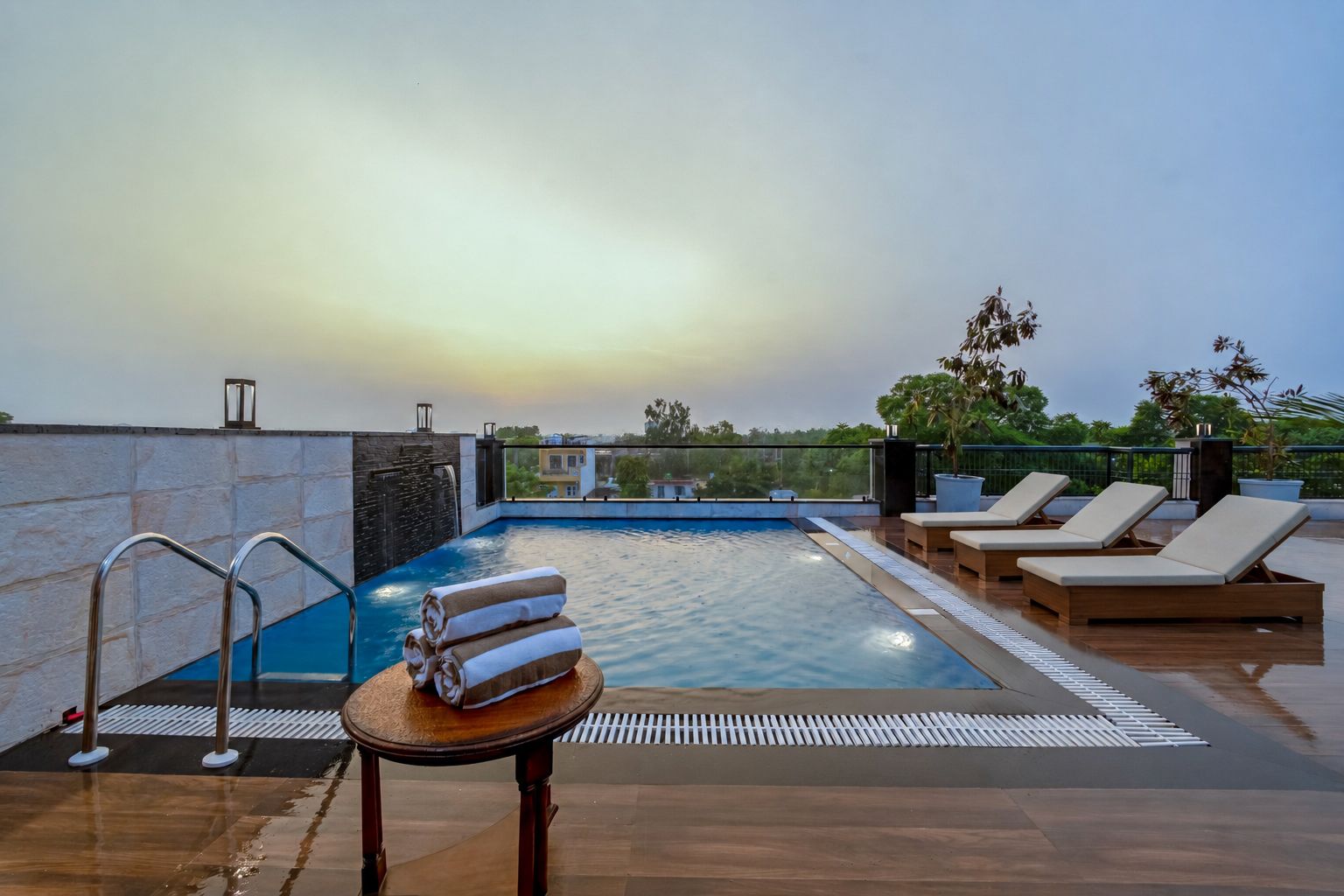 Un área de piscina en la azotea con sillones, una pequeña mesa con toallas y vistas panorámicas de la ciudad.
