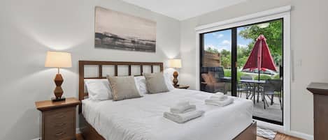 A hotel room with a large bed, two bedside tables with lamps, a painting on the wall, and a sliding glass door leading to an outdoor area with a red umbrella.