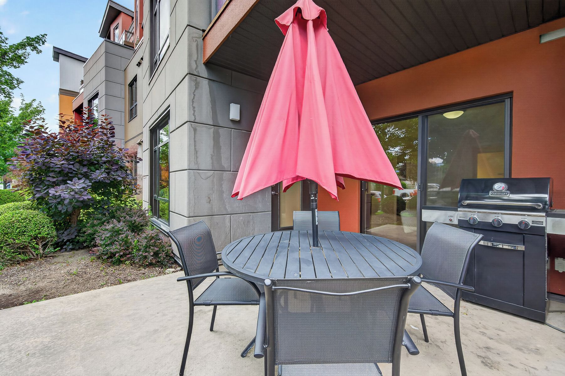 An outdoor patio with a round table and two chairs, a red umbrella, and a barbecue grill.