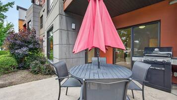 An outdoor patio with a round table and two chairs, a red umbrella, and a barbecue grill.