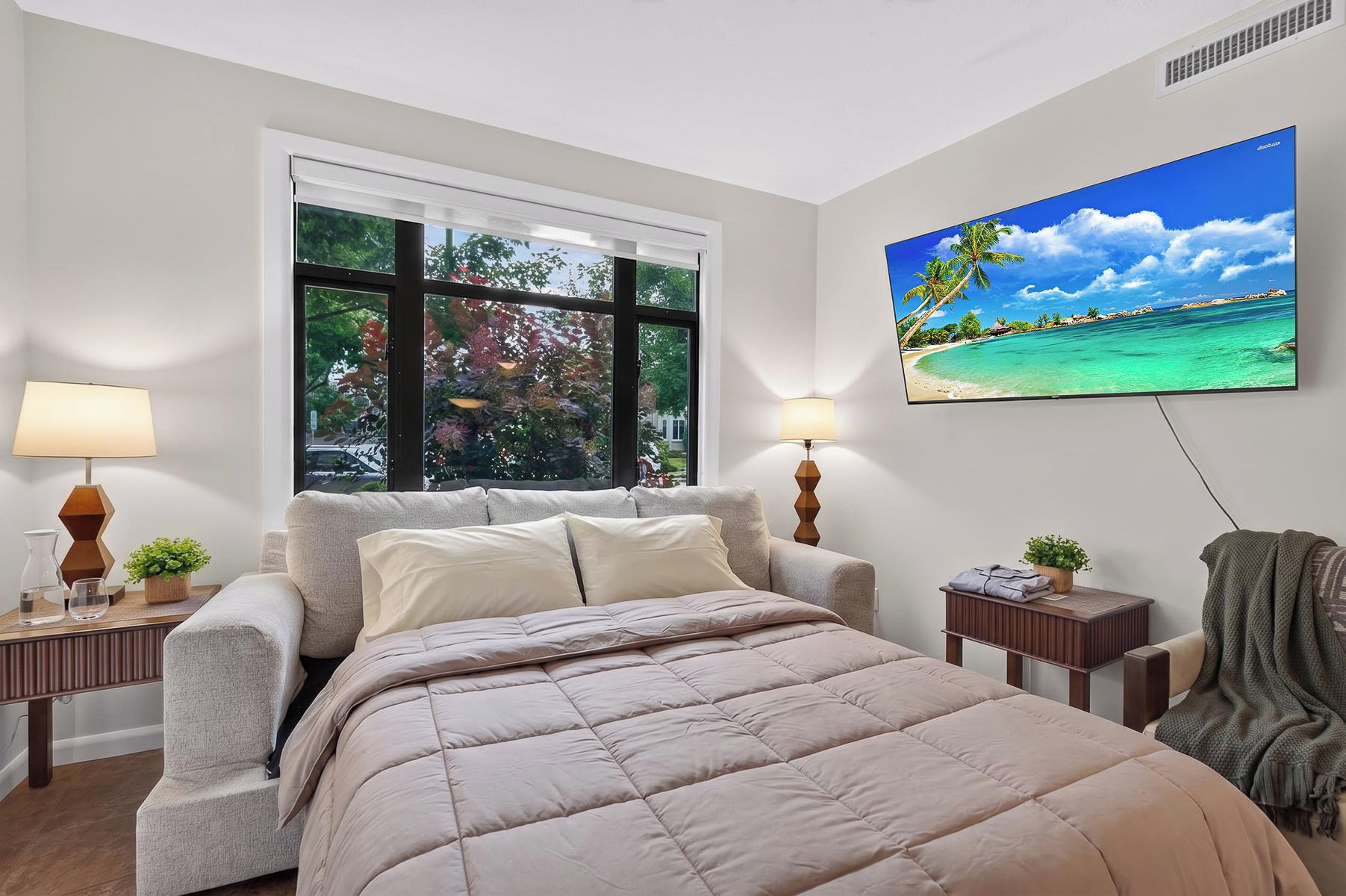 Un dormitorio con cama, ventana, televisor montado en la pared y mesita de noche con lámpara.
