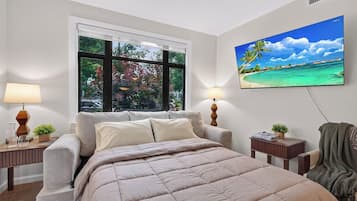 Un dormitorio con cama, ventana, televisor montado en la pared y mesita de noche con lámpara.