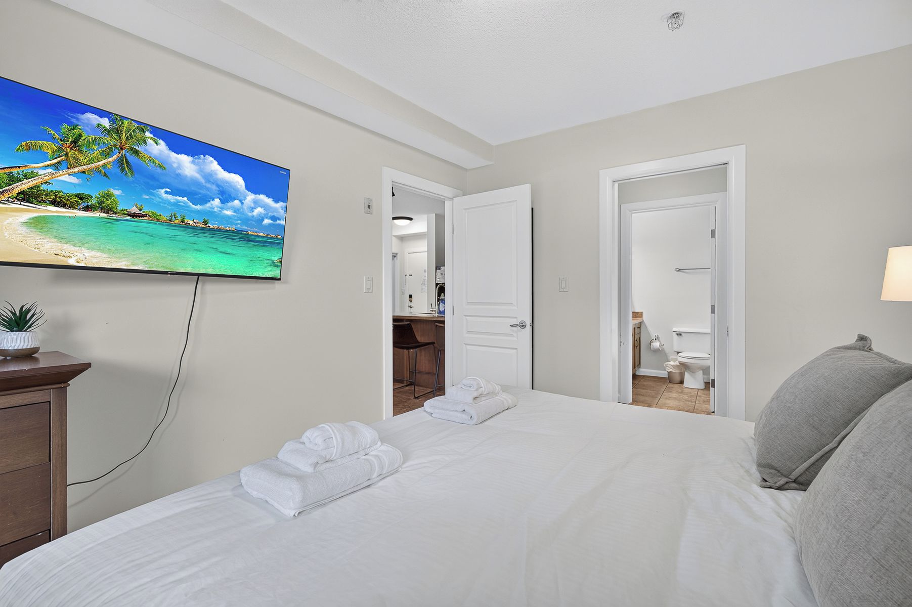 A hotel room with a bed, a TV displaying a beach scene, a desk with a chair, and a bathroom visible through an open door.
