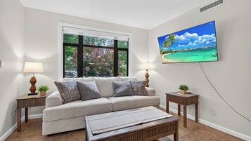 A living room with a sofa, two side tables with lamps, a coffee table, a large window with a view of trees, and a wall-mounted TV displaying a tropical beach scene.