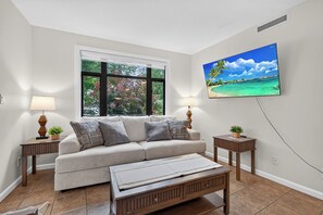 A living room with a sofa, two side tables with lamps, a coffee table, a large window with a view of trees, and a wall-mounted TV displaying a tropical beach scene.