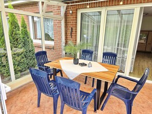 Eine Terrasse im Freien mit einem Holztisch, blauen Plastikstühlen, einem Blumentopf und einer Ziegelwand.
