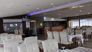 A restaurant with white upholstered chairs, tables set with plates and glasses, and a bar area in the background. - GRAND ALİN HOTEL (Turhal)