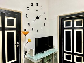 Ein Zimmer mit einer Wanduhr, einem Fernseher auf einem Glastisch und schwarzen Türen mit weißem Rahmen.
