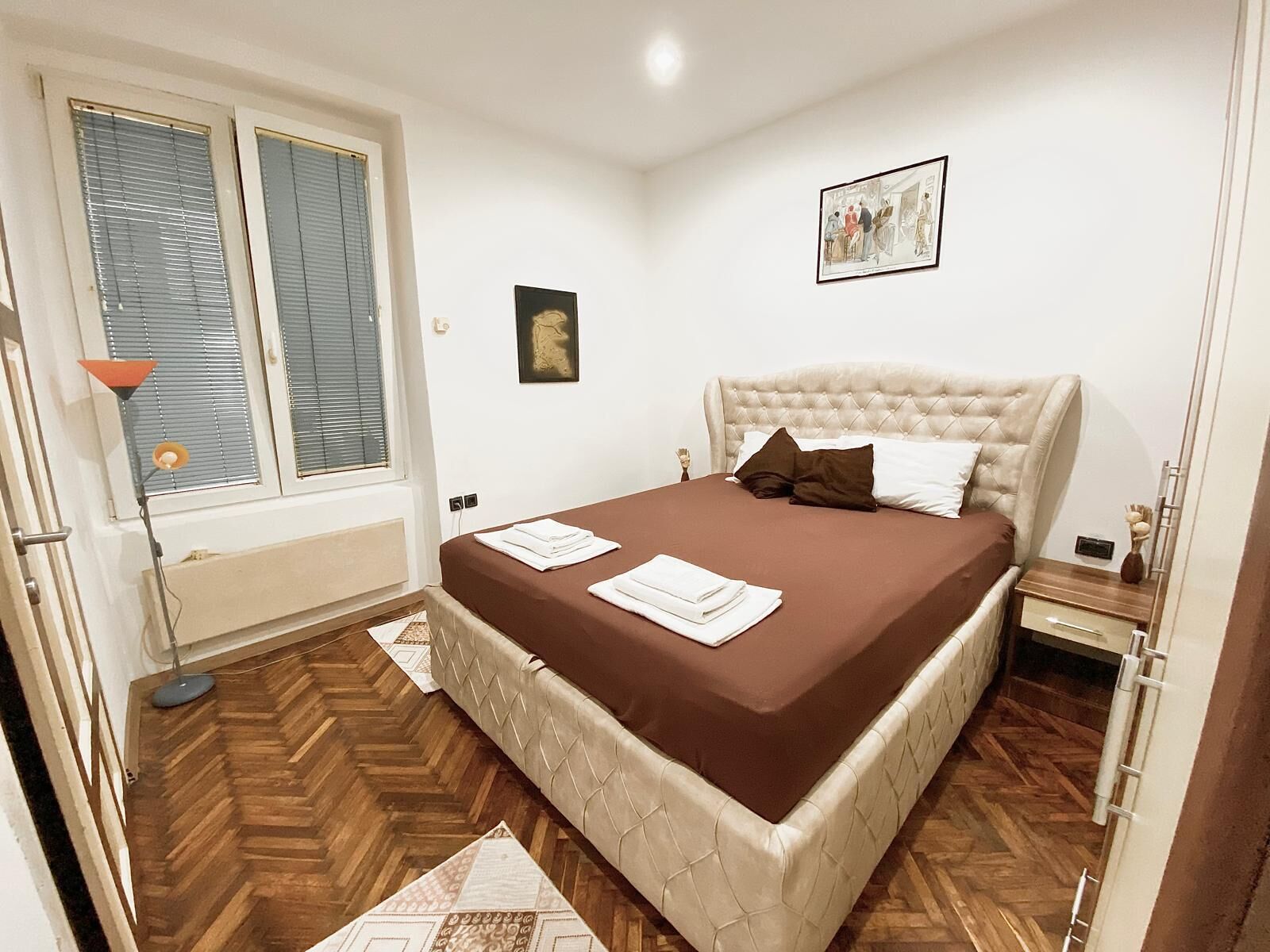 Habitación de hotel con una cama grande, una mesita de noche, una lámpara de pie y una ventana con persianas.
