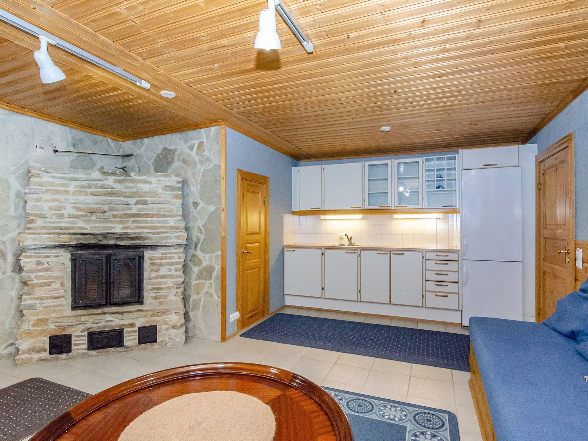 A room with a stone fireplace, wooden ceiling, a kitchen with white cabinets, and a blue sofa.
