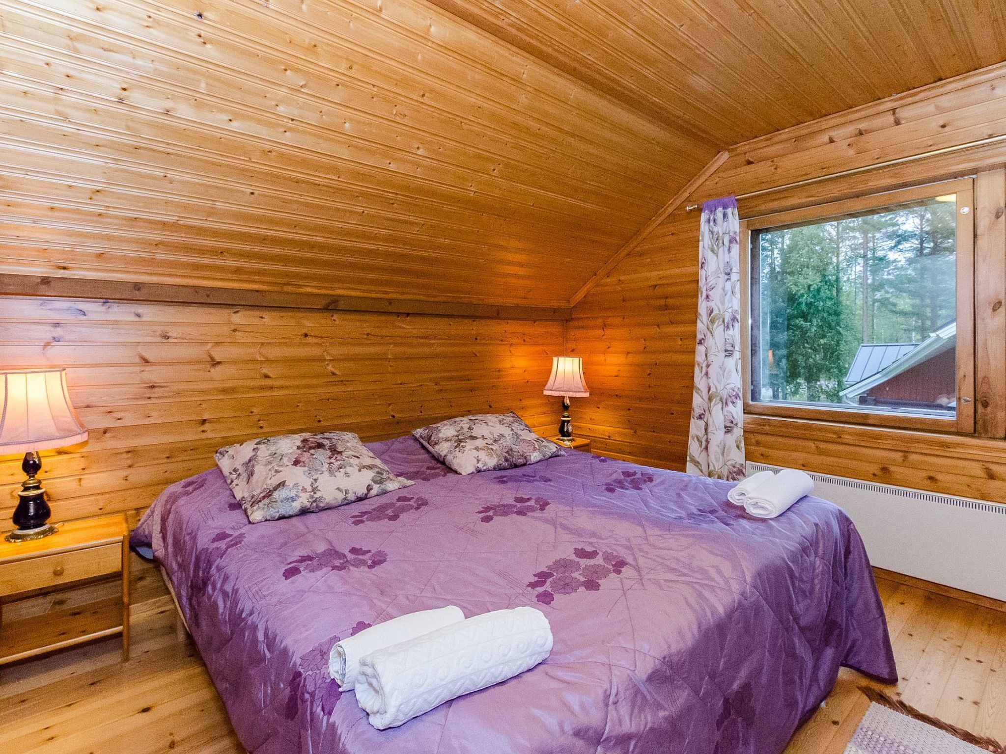 Een knus slaapkamer in een houten hutje, met een bed, twee bedlampen en een raam met uitzicht op bomen.