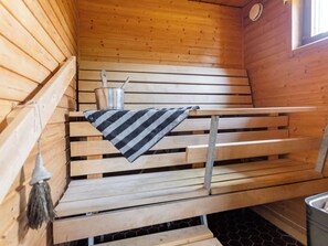 Banco de sauna de madeira com toalha listrada, balde de metal com concha e uma vassoura pendurada na parede.